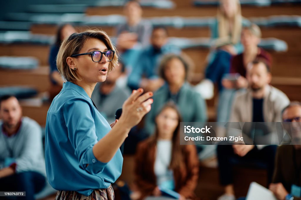 Mid adult businesswoman talking to group of seminar attendees in conference hall.